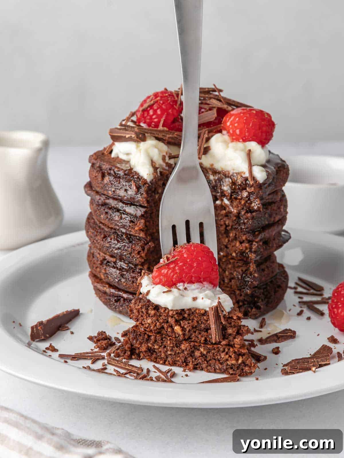 A fork lifts a bite-sized piece of chocolate protein pancake, revealing its fluffy interior, topped with fresh raspberries and a hint of syrup.