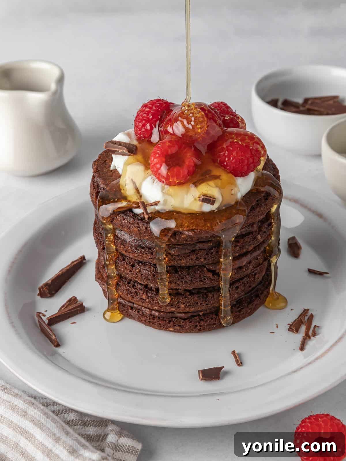 Golden maple syrup being generously drizzled over a stack of warm chocolate protein pancakes on a white plate, highlighting their appetizing texture and inviting rich color.