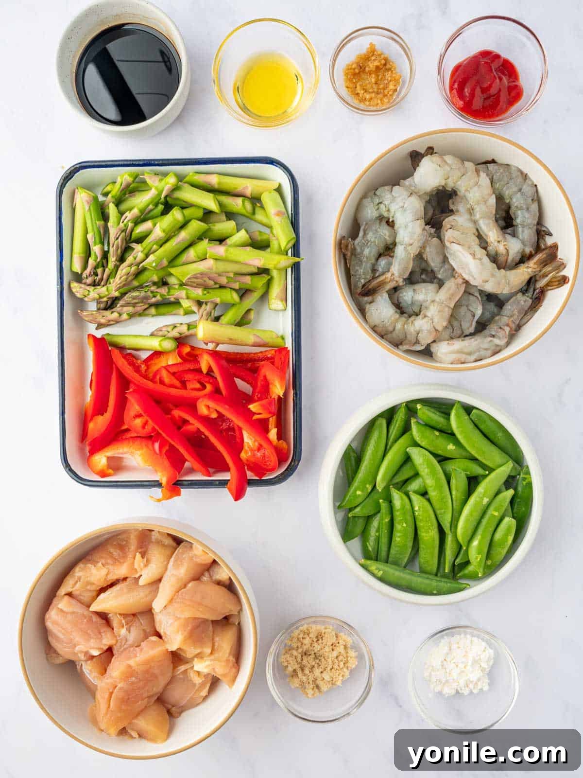 Sizzling Surf and Turf Stir-Fry 3 A collection of fresh ingredients arranged on a counter, including raw chicken pieces, peeled shrimp, sliced red bell peppers, asparagus, snow peas, and small bowls containing ginger, garlic, soy sauce, and sambal oelek, all ready for the stir fry.