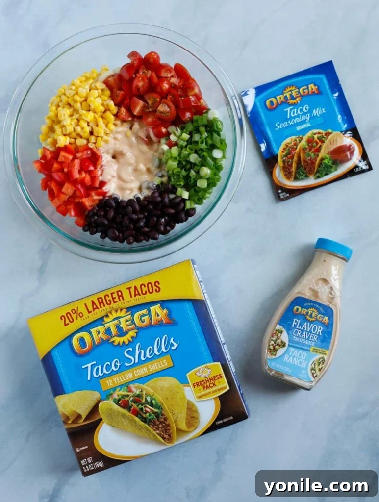 A vibrant selection of fresh ingredients for Taco Ranch Pasta Salad, including elbow pasta, grape tomatoes, corn, black beans, green onions, red bell pepper, lime, seasoning, and cheese, displayed on a kitchen counter.