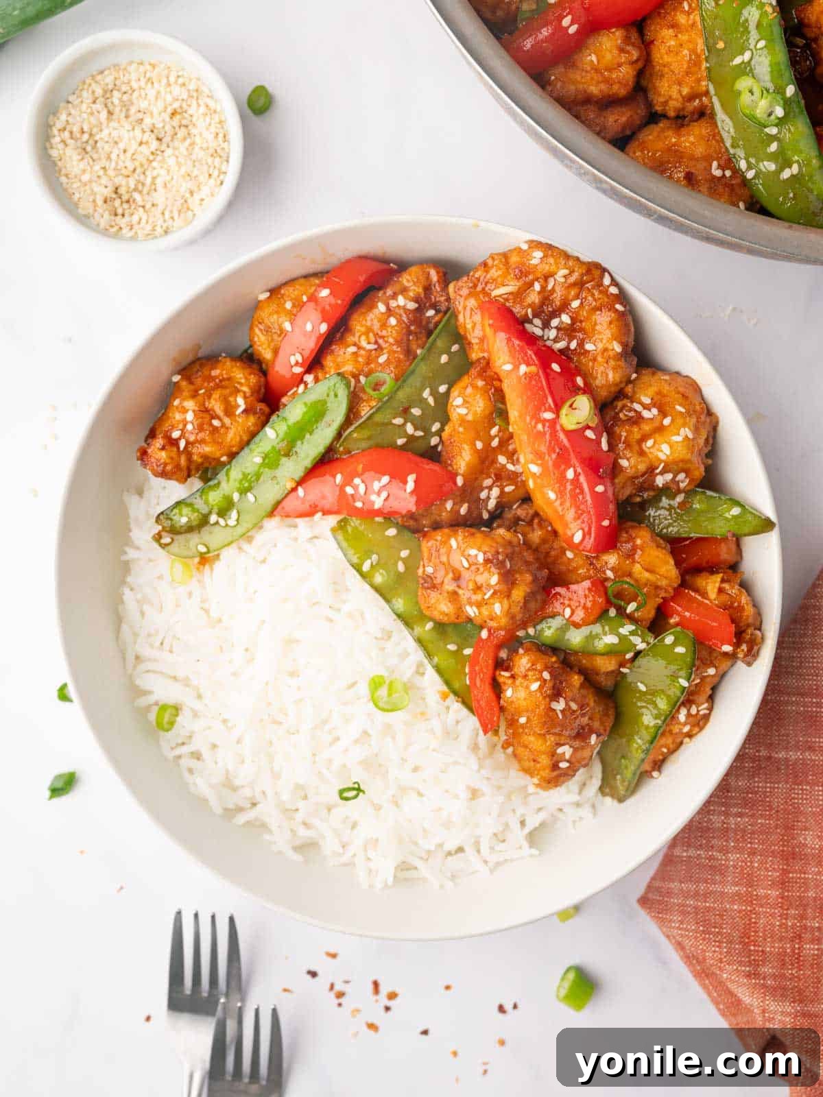 A plate with rice, veggies and crispy honey chili chicken garnished with sesame seeds.