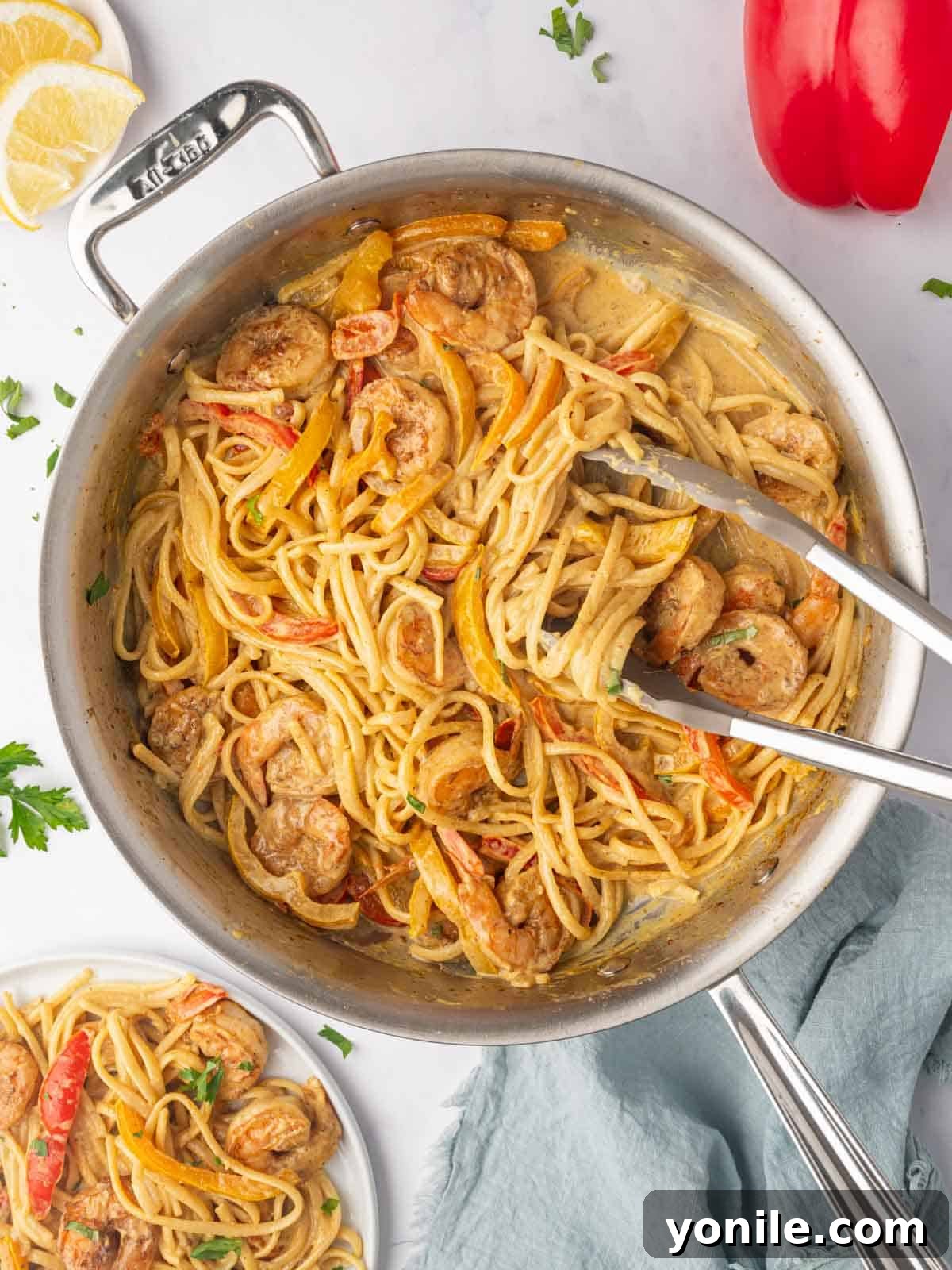 Serving tongs expertly scoop out a generous portion of Jamaican Rasta Pasta with succulent shrimp from a simmering skillet, showcasing its vibrant appeal.