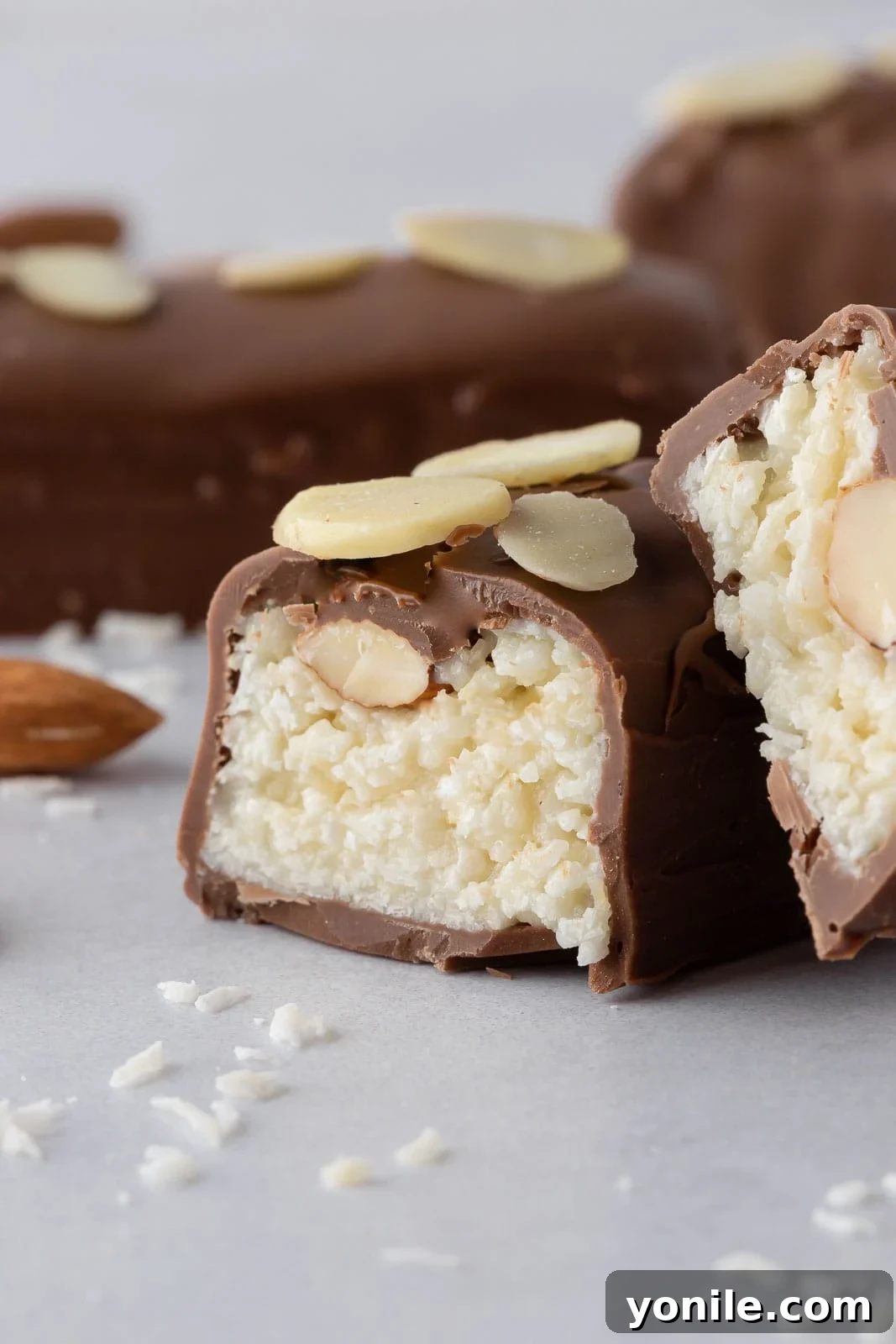 A close-up of a homemade coconut chocolate bar, cut in half to reveal the moist, chewy coconut and almond filling, encased in a rich chocolate shell.