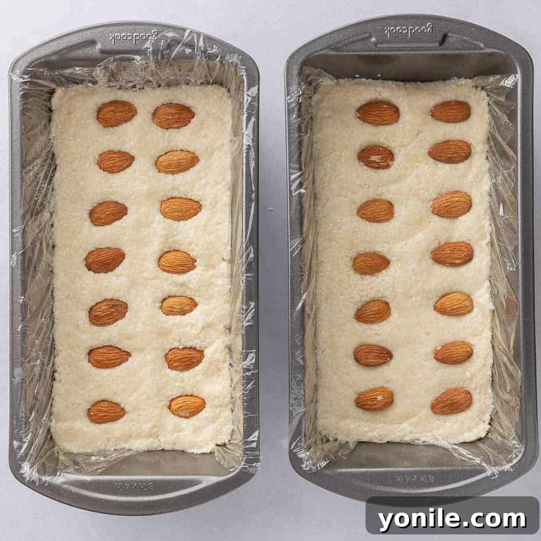 Coconut and almond candy bar filling is pressed into two pans lined with plastic wrap, demonstrating the setting process for the no-bake bars before chilling.