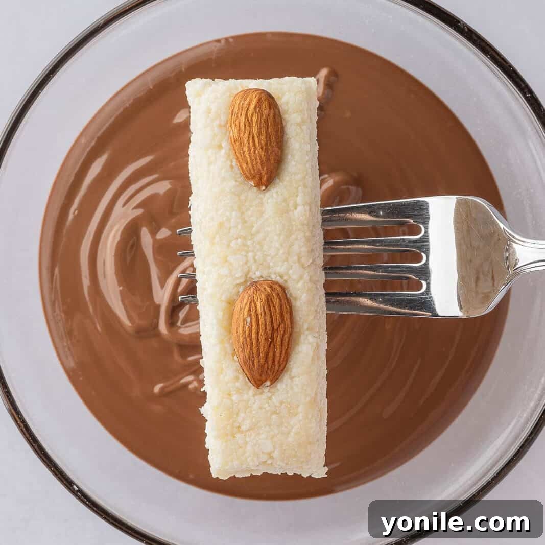 A bowl of glossy, melted chocolate is shown, representing the smooth coating ready for dipping the homemade coconut almond bars.