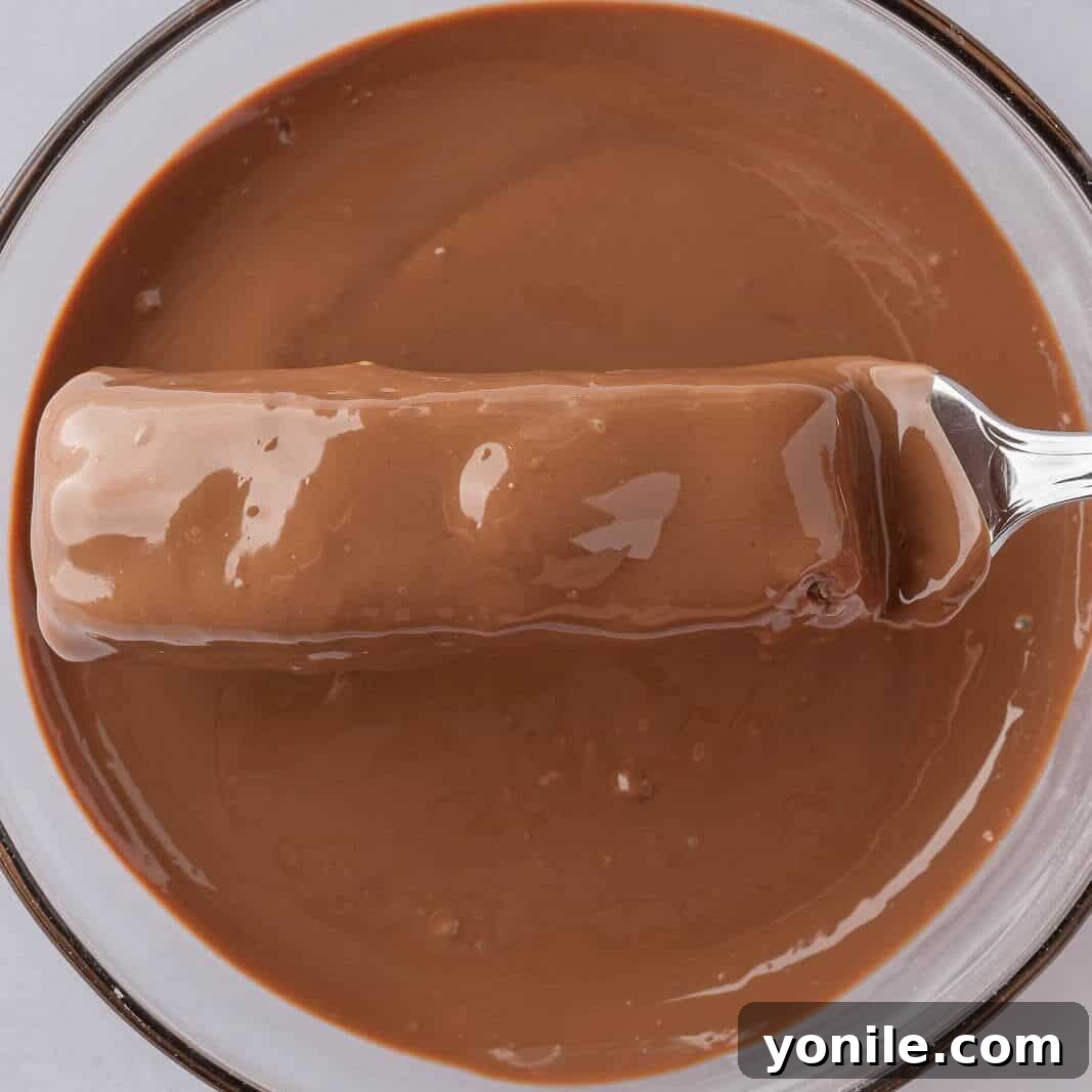 A homemade coconut bar is carefully dipped into a bowl of melted chocolate using a fork, ensuring an even and complete coating for the delicious treat.