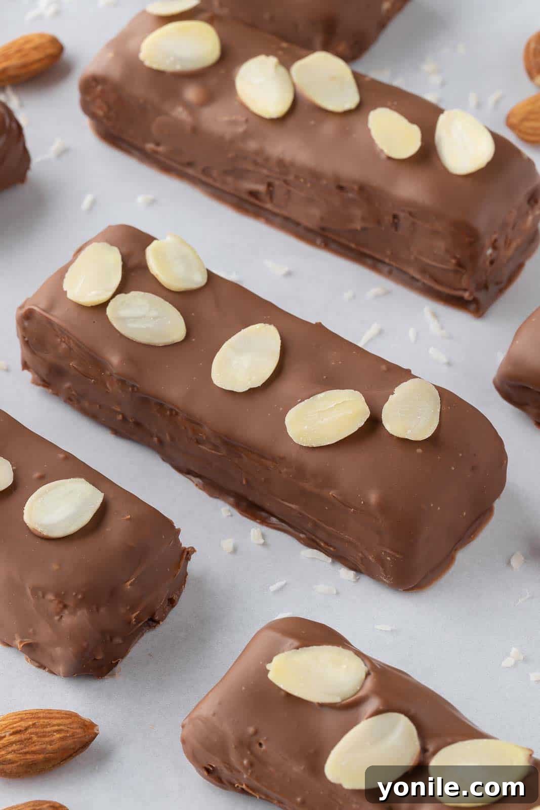 A close-up view of three homemade coconut chocolate bars, garnished with slivered almonds, displayed on a tray against a white background.