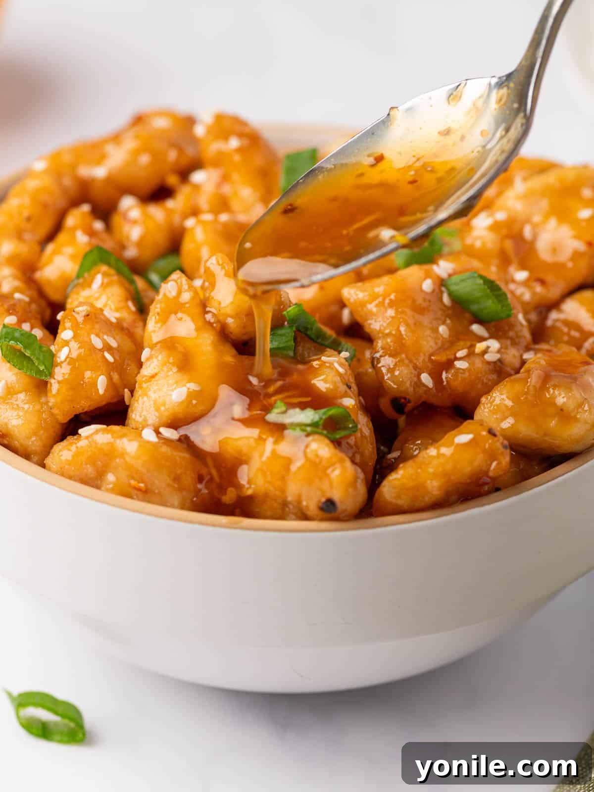 A spoon generously drizzles homemade orange chicken sauce over a bowl of perfectly fried chicken pieces, ready to be enjoyed.