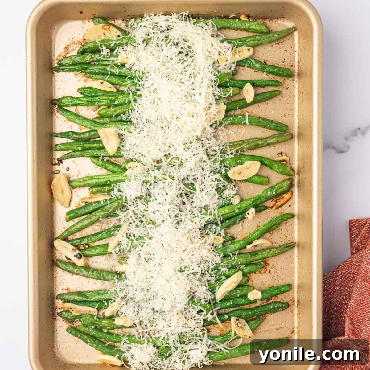 Freshly grated parmesan cheese being sprinkled over baked green beans on a tray.
