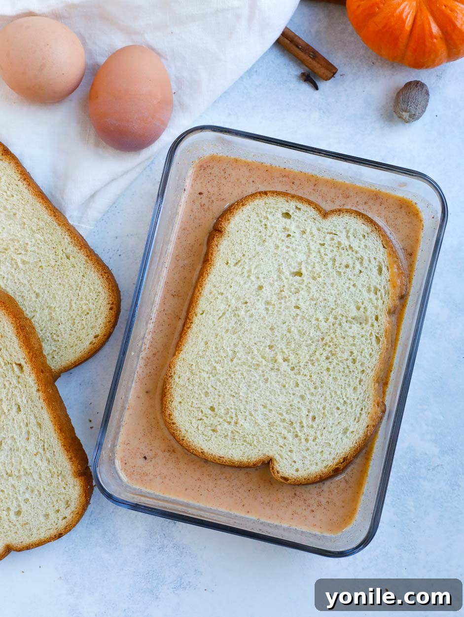 Dipping Brioche for French Toast A slice of brioche bread being generously dredged in a rich pumpkin spice egg mixture in a glass bowl.