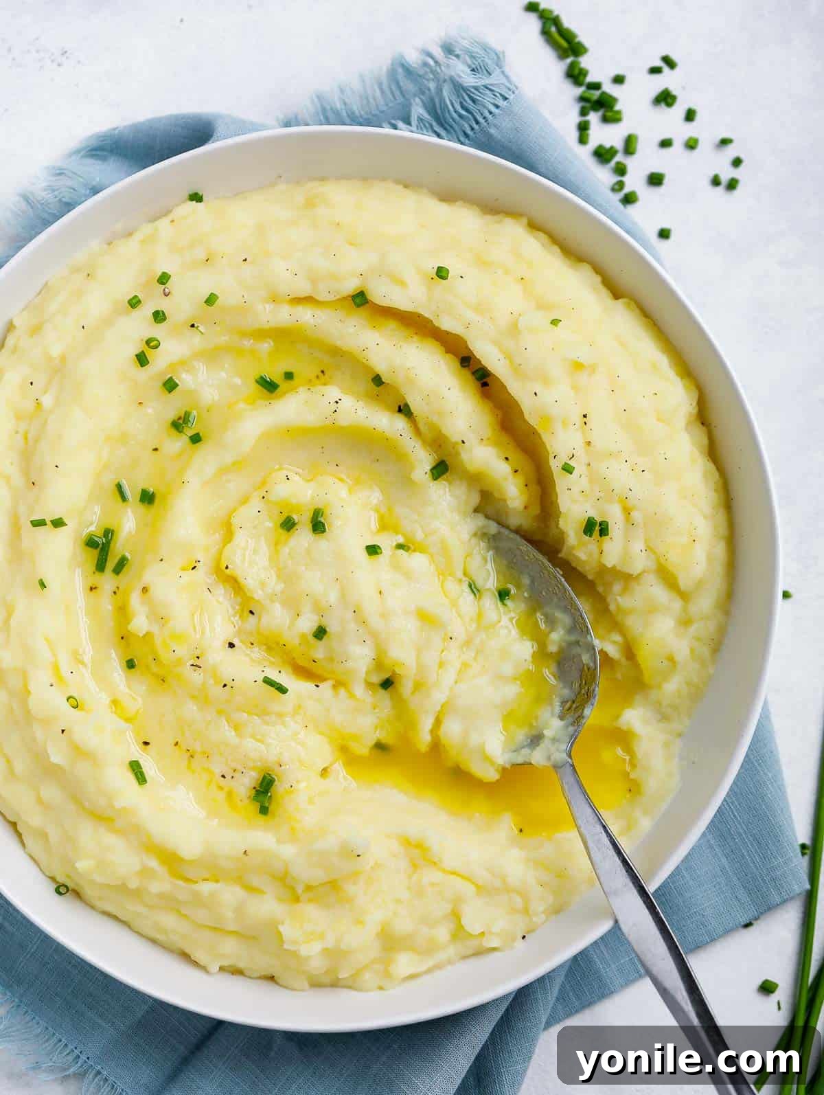 Easy Homemade Mashed Potatoes creamy mashed potatoes in a white plate topped with melted butter and chives, with a spoon in the plate