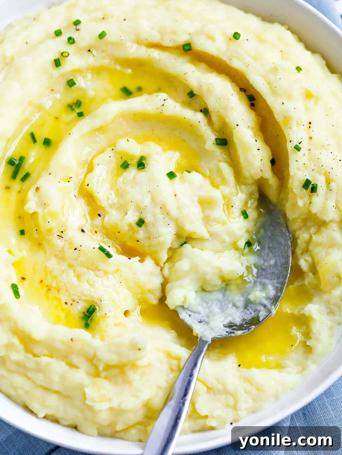 Effortless Velvety Mash 4 creamy mashed potatoes in a white plate topped with melted butter and chives, with a spoon in the plate