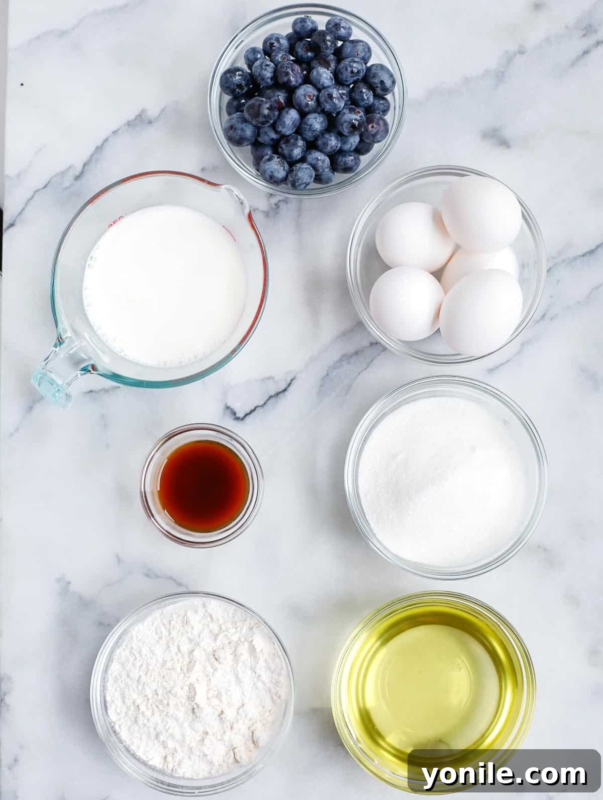 Luscious Blueberry Bundt Cake 2 A selection of fresh ingredients including flour, eggs, milk, sugar, oil, baking powder, vanilla extract, salt, fresh blueberries, and lemon zest laid out on a table.