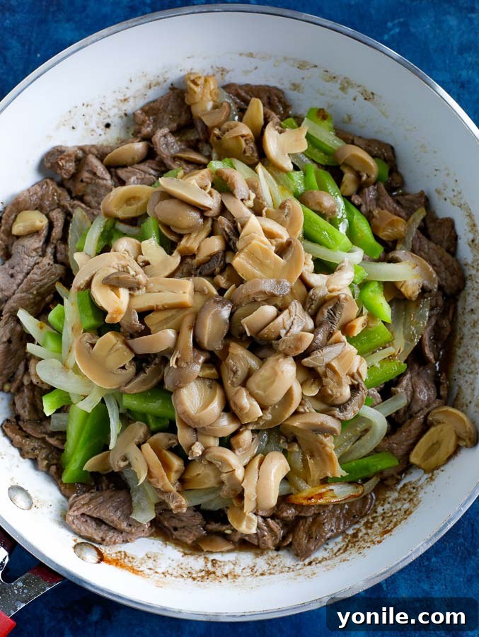 Steak, peppers, onions, and mushrooms cooking in a non-stick skillet on a blue counter, combining flavors.