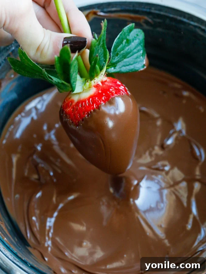 A fresh strawberry being carefully dipped into a bowl of perfectly melted, glossy chocolate, ensuring even coverage.