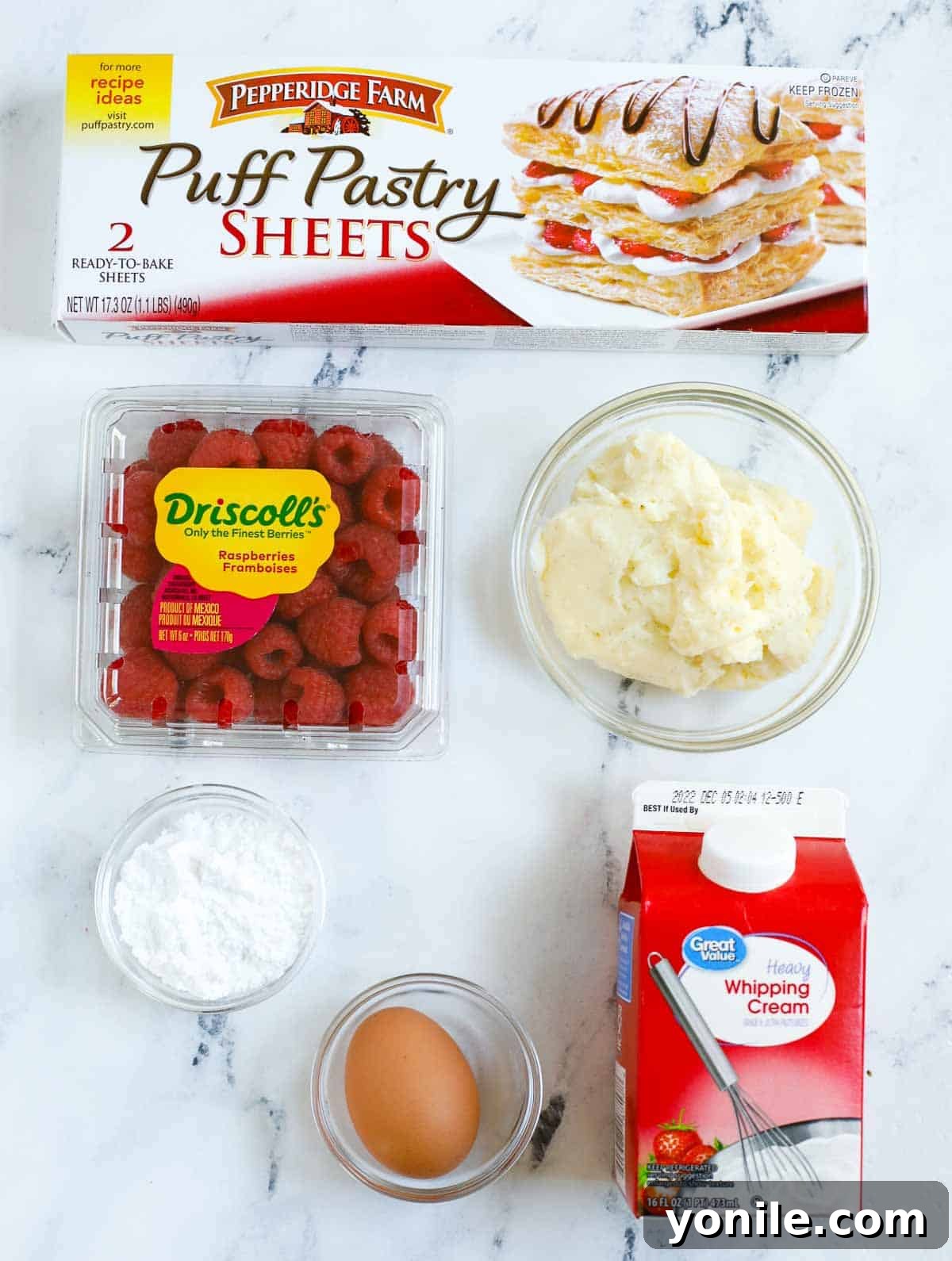 A beautiful flat lay of fresh raspberry tart ingredients: puff pastry, fresh raspberries, egg, powdered sugar, and a bowl of pastry cream.