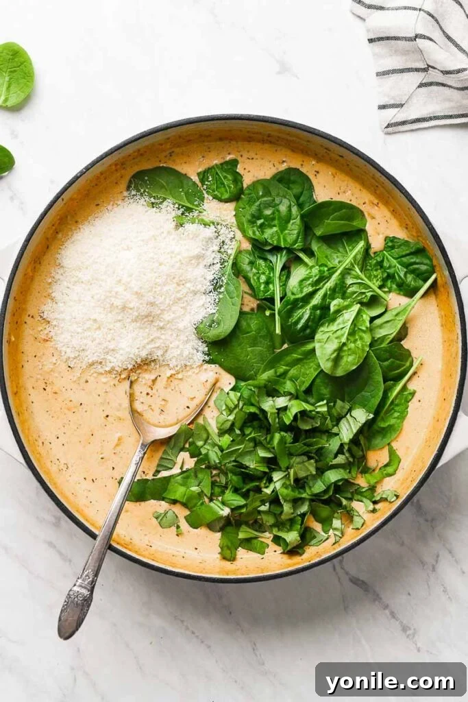 Velvety Sun-Dried Tomato Chicken Thighs 4 Parmesan cheese and fresh baby spinach being added to the creamy sauce in a skillet.