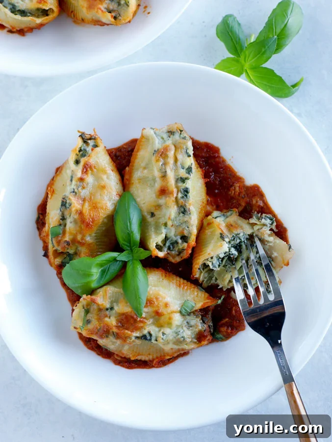 A white bowl with 4 spinach artichoke stuffed shells with a fork and some fresh basil.