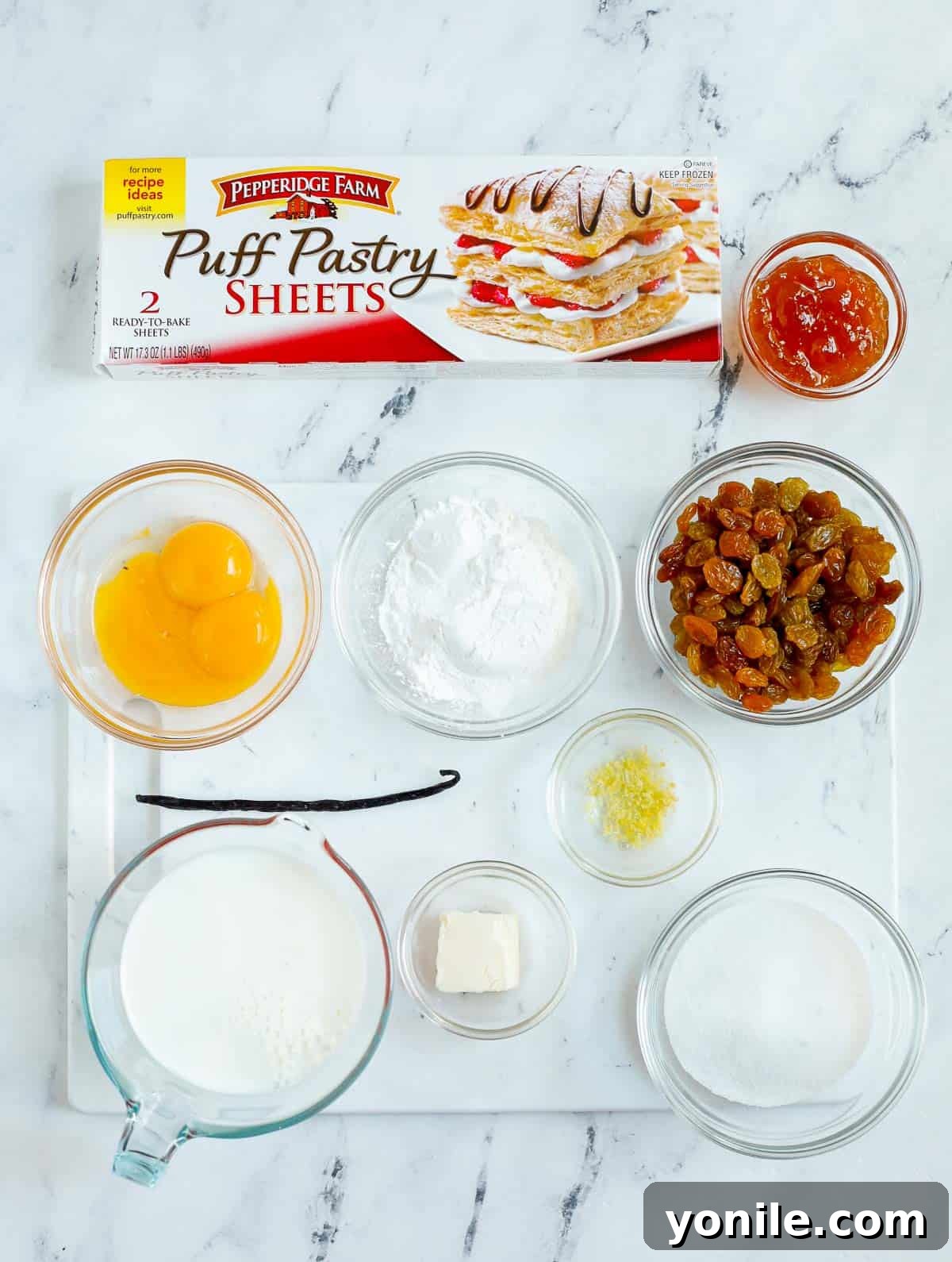 All the necessary ingredients for making pain aux raisins laid out on a white surface, including puff pastry, raisins, apricot preserve, and the components for crème patissiere.