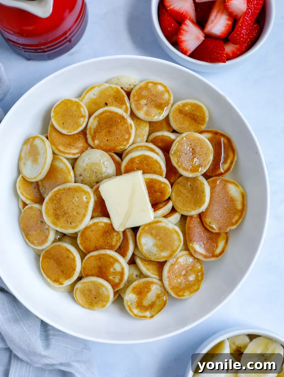 A bowl of mini pancake cereal with a dollop of butter and a drizzle of syrup, garnished with fresh banana slices and strawberries.