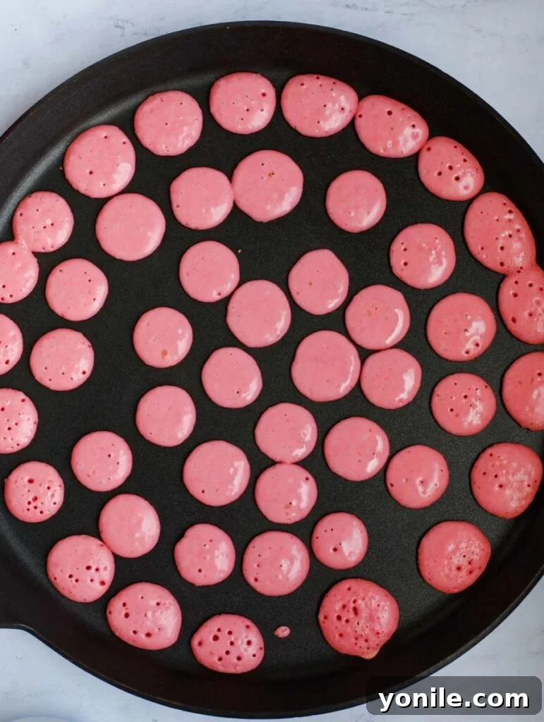 Mini strawberry pancakes cooking on a pan, displaying their charming pink hue.