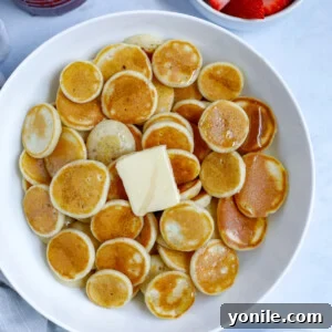 A bowl of mini pancake cereal with a dollop of butter and a drizzle of syrup, garnished with fresh banana slices and strawberries.