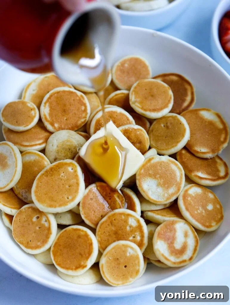 Pancake syrup being generously poured over a bowl of freshly cooked mini pancakes, enhancing their golden appeal.