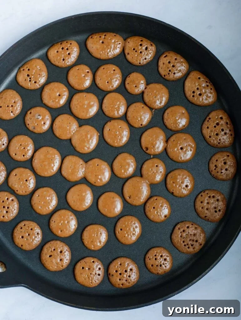 Mini chocolate pancakes cooking on a pan, showing their dark, appealing hue.