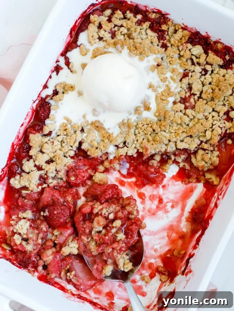 Freshly baked strawberry crisp in a baking dish, with a serving spoon and a scoop of vanilla ice cream, ready to be enjoyed.