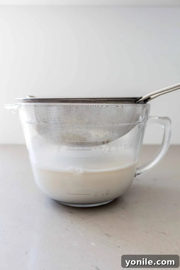 Homemade oat milk being strained through a fine-mesh strainer into a glass bowl