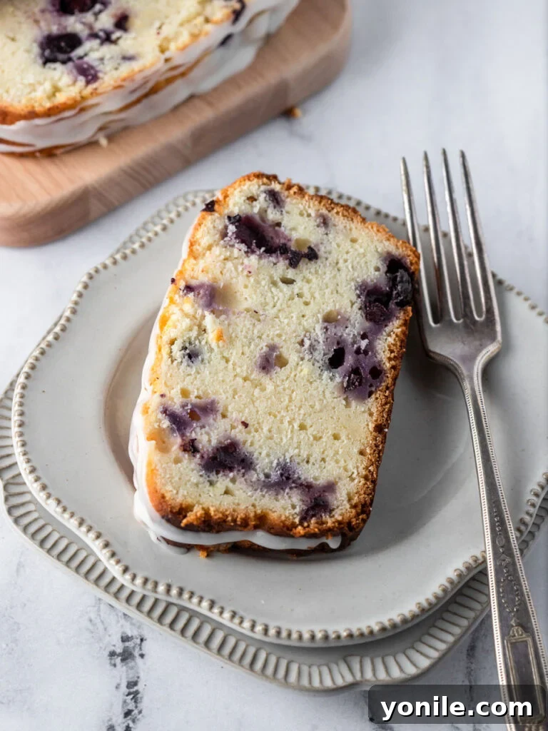 Bright Lemon Blueberry Bread 6 A single slice of moist Lemon Blueberry Bread, glistening with lemon glaze, neatly placed on a pristine white plate.