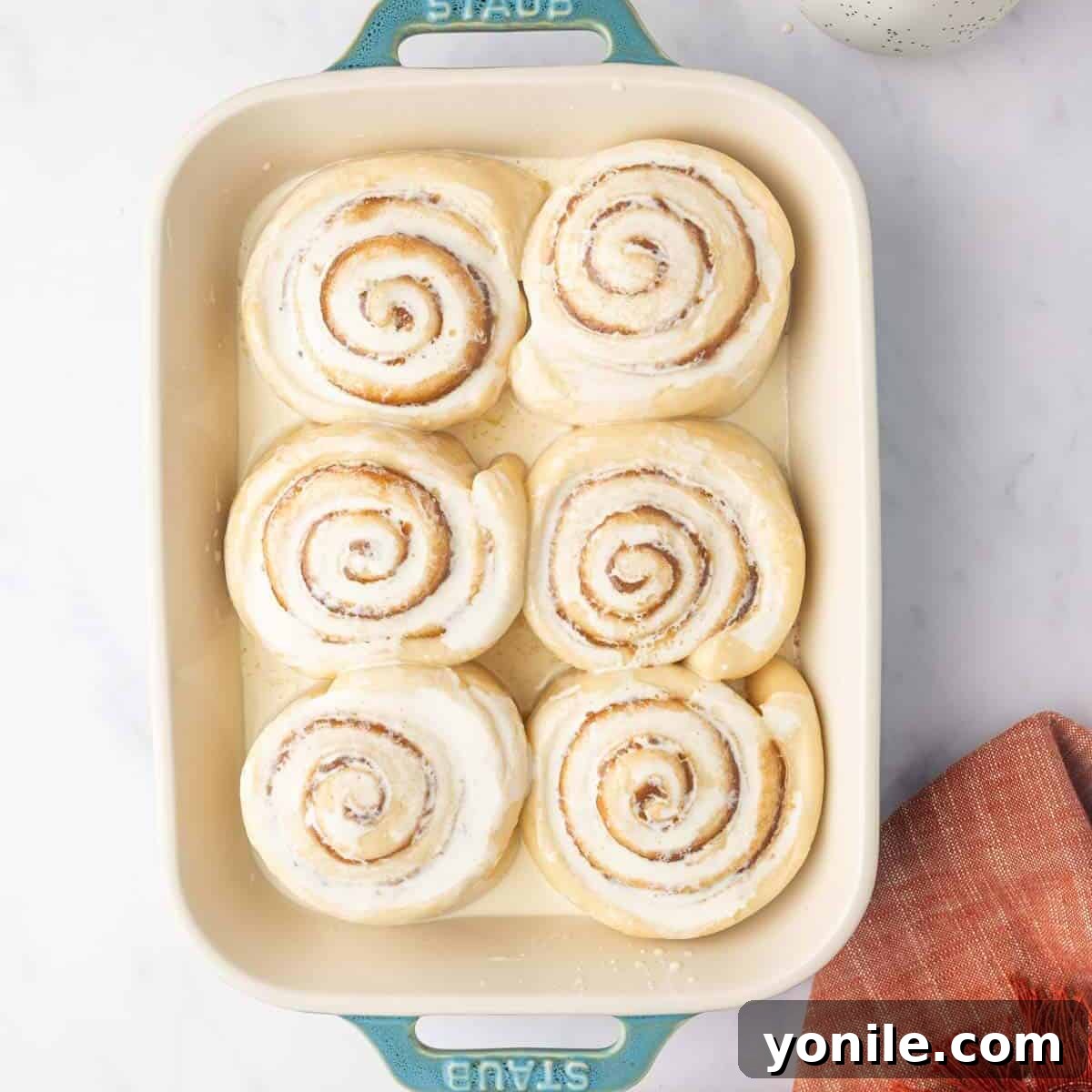 Warm heavy cream is poured evenly over the unbaked, risen cinnamon rolls in a rectangular baking dish, preparing them for baking.
