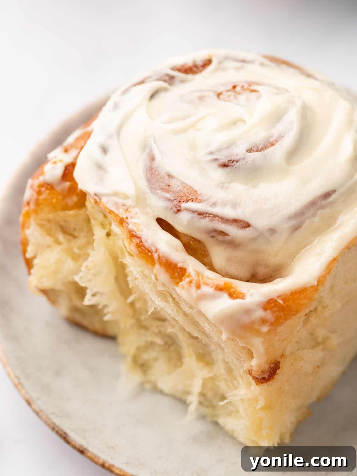 Close-up of a soft, flaky cinnamon roll generously topped with cream cheese frosting, served on a white plate, ready to be enjoyed.