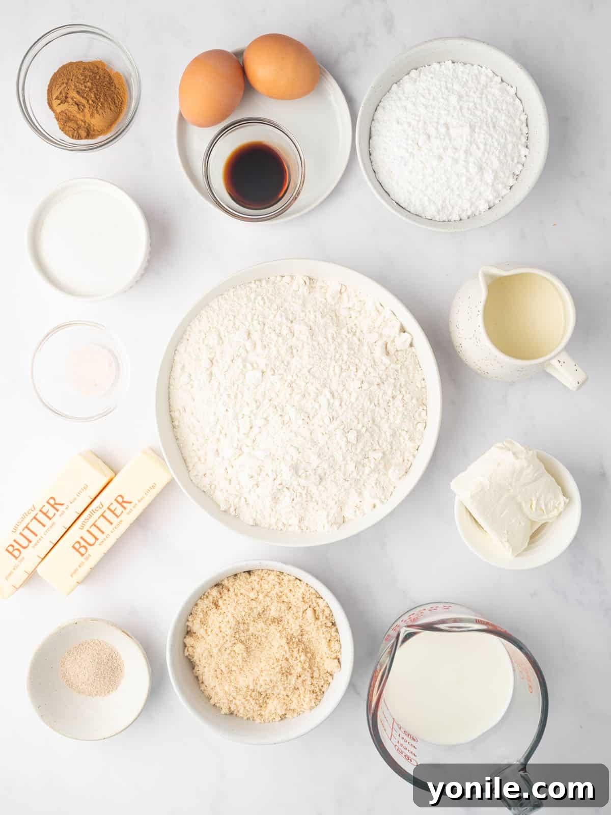 A collection of ingredients laid out, including flour, sugar, yeast, milk, butter, eggs, vanilla, heavy cream, brown sugar, cinnamon, and confectioners' sugar, ready for making heavy cream cinnamon rolls.