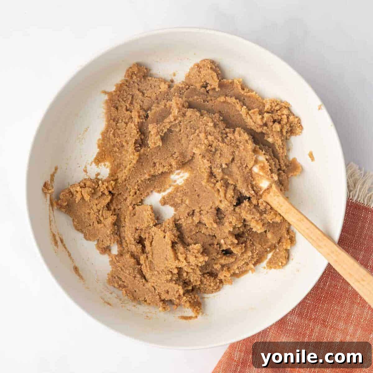 Cinnamon roll filling, a mixture of butter, brown sugar, salt, and cinnamon, is mixed until smooth in a white bowl with a wooden spoon.