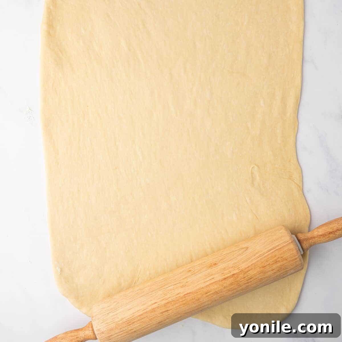 The risen cinnamon roll dough is rolled into a large, even rectangle on a lightly floured countertop using a wooden rolling pin.