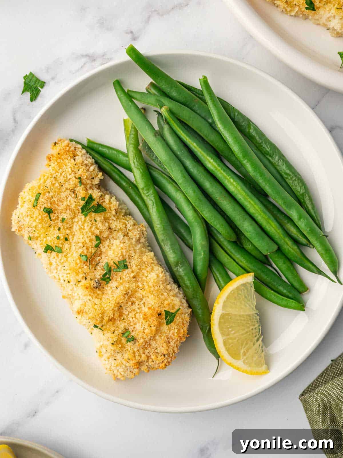 Baked cod on a plate with green beans, garnished with fresh herbs and lemon.